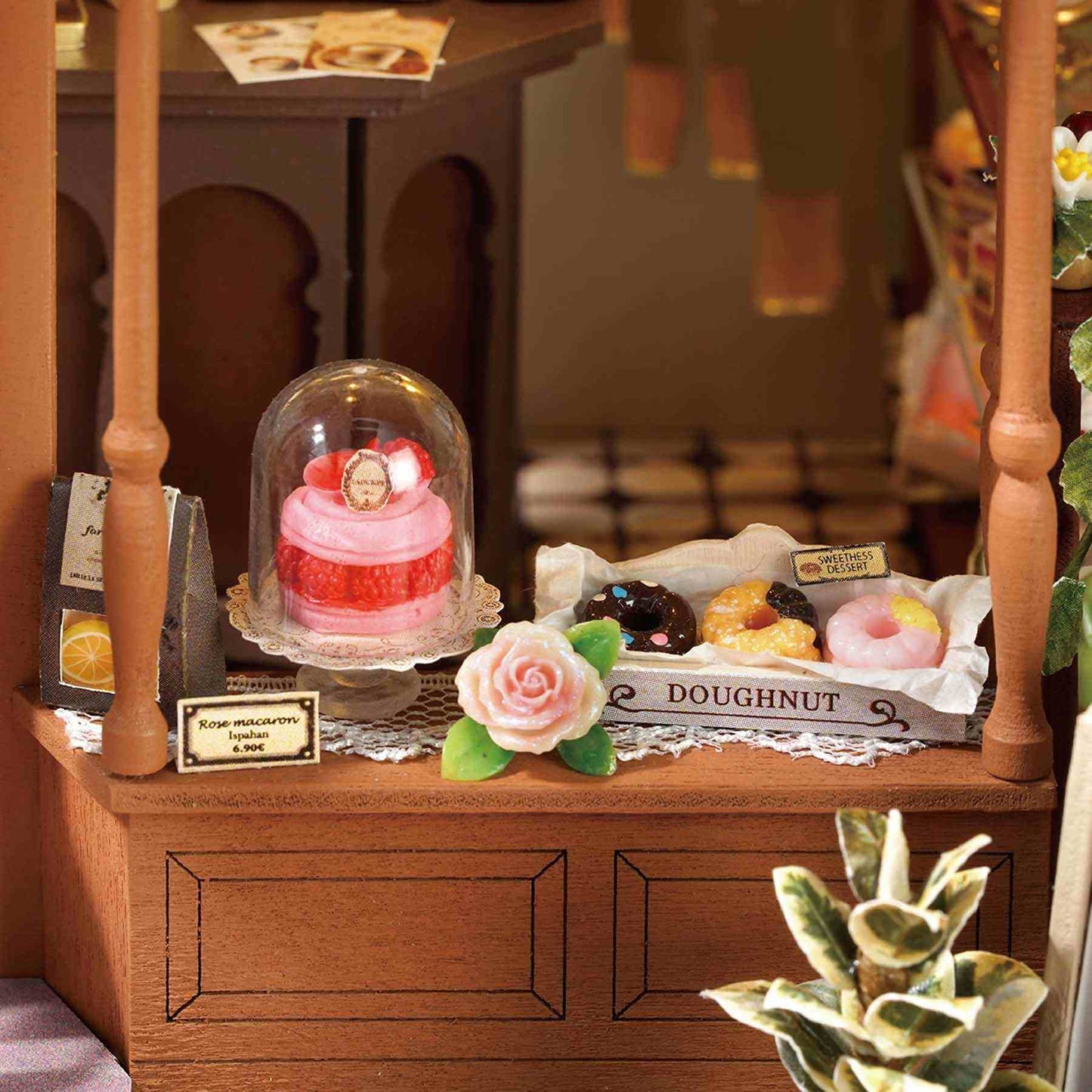 Miniature bakery display with a pink cake under a glass dome on a wooden counter.