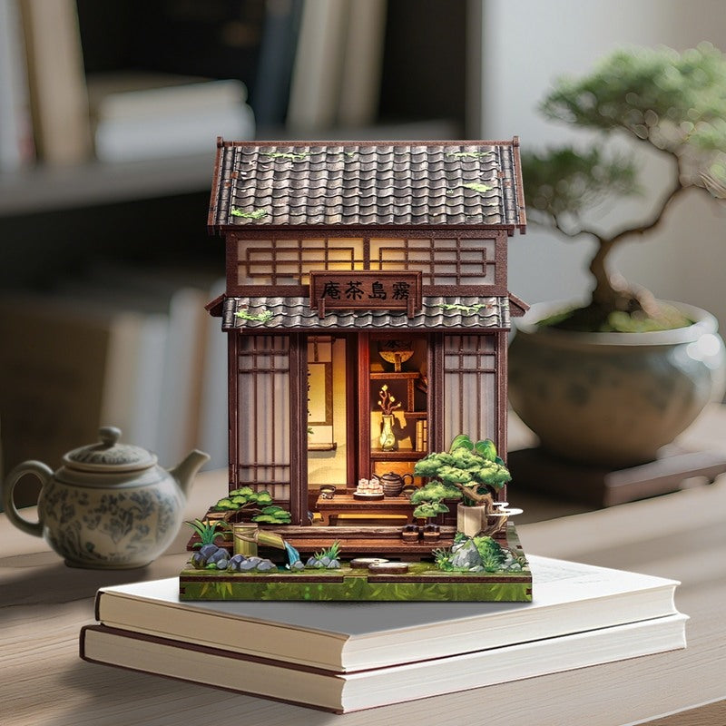 Miniature model of a traditional Japanese building on a table with a teapot and bonsai tree in the background.