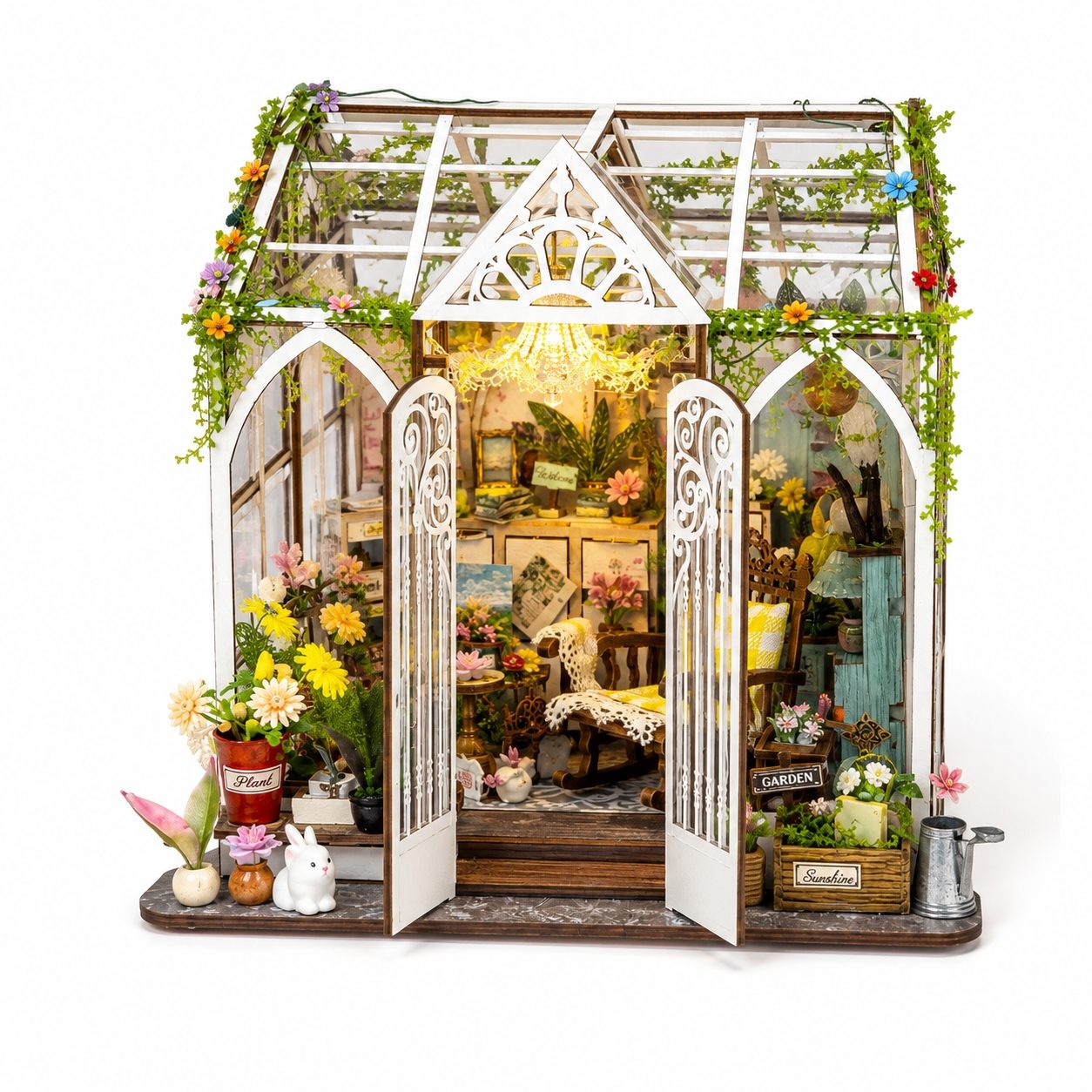 A miniature greenhouse dollhouse with white arched frames, glass panels, and lush green vines draped along the roof and walls. The open doors reveal a detailed, cozy interior filled with tiny furniture pieces, potted plants, and warm glowing lights.
