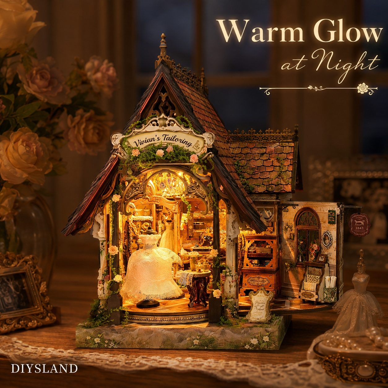 A miniature dollhouse-style shop set labeled 'Vivian's Tailoring' with warm internal lighting, displayed on a tabletop in a softly lit room. The scene includes decorative flowers, lace, and the text 'Warm Glow at Night'.