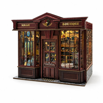 A detailed miniature model of a wizarding shop front, labeled 'MAGIC BOUTIQUE' with a Slytherin crest, featuring dark reddish-brown wood paneling, glass windows, and warm yellow interior lighting, shown on a white background.