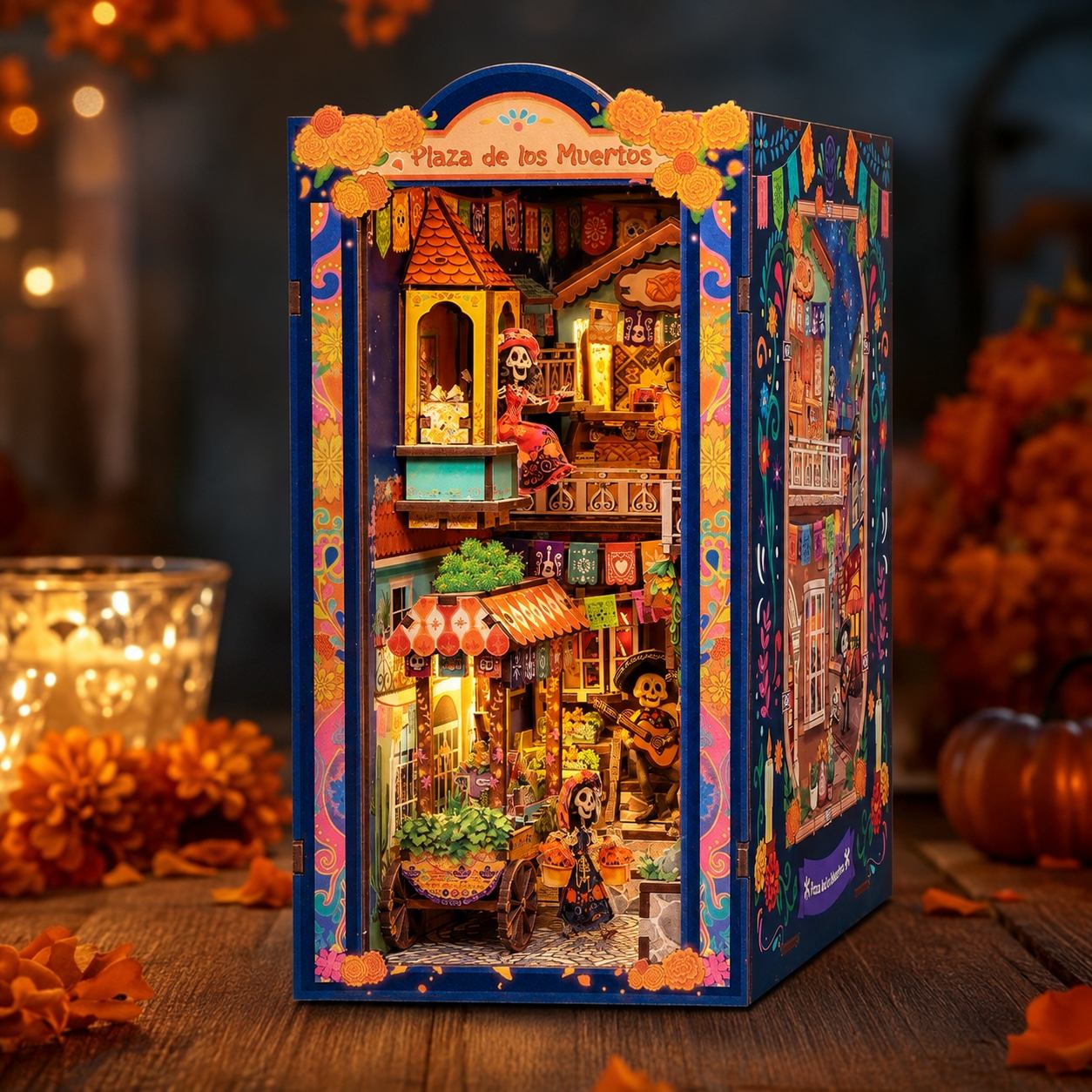 A Dia de los Muertos themed miniature book nook with colorful street details, glowing windows, and small figurines, displayed on a wooden surface with warm autumn decor in the background.