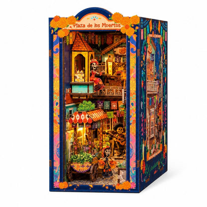 A colorful miniature Dia de los Muertos (Day of the Dead) city book nook model titled 'Plaza de los Muertos', featuring a detailed multi-story street scene with skeleton characters, orange marigolds, and traditional Mexican architecture, shown on a clean white background.