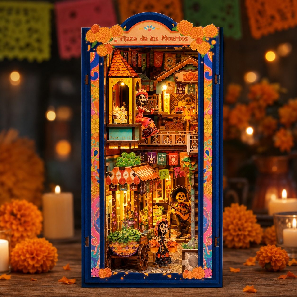 A decorative miniature diorama titled 'Plaza de los Muertos' stands centered on a table, surrounded by lit candles, orange marigolds, and a warm, blurred festive background. The structure features a blue frame with colorful floral and scroll borders and a detailed interior scene with tiny houses, candles, and skeleton figures.