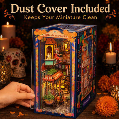 A hand holds a clear dust cover in front of a colorful miniature Dia de los Muertos city building kit box labeled 'Plaza de los Muertos.' The box shows a detailed multicolor diorama with glowing windows, candles, flowers, and skeleton figures.