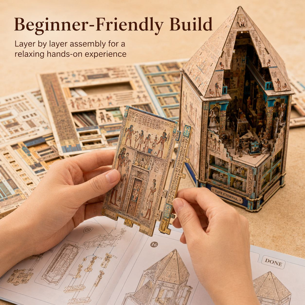 A person's hands are shown assembling a wooden 3D model kit, holding a wooden panel with ancient Egyptian-style illustrations. In the background, a completed multi-level house model is displayed next to its instruction sheets on a beige surface.