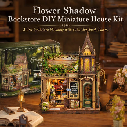 Flower Shadow Bookstore DIY Miniature House Kit displayed on a wooden table with its packaging box in the background. The miniature house features a detailed Victorian-style bookstore with warm yellow glowing windows, green foliage accents, and a small lantern on the porch.