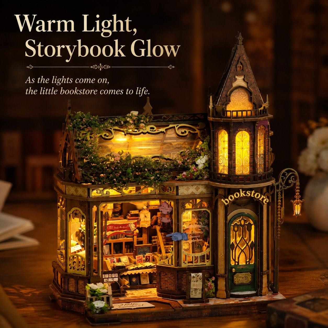 A miniature warm-white lit bookstore dollhouse or model building displayed on a wooden surface, with glowing yellow windows, greenery details, and decorative storefront elements. Text in the upper left reads, 'Warm Light, Storybook Glow' and 'As the lights come on, the little bookstore comes to life.