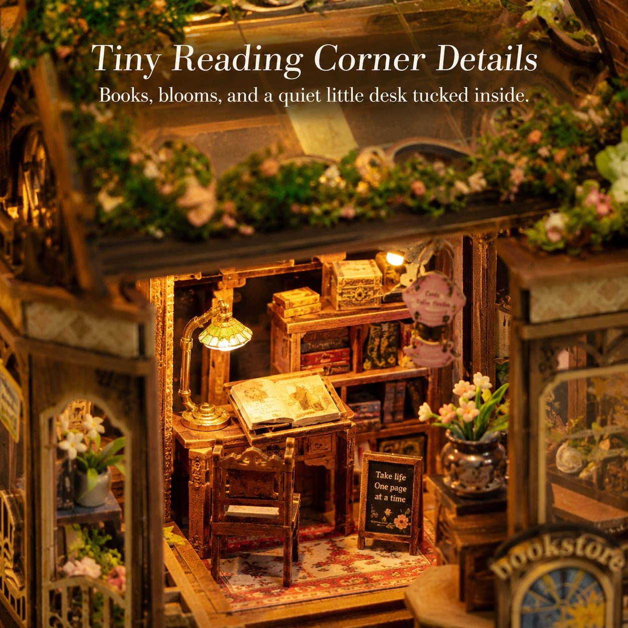 A miniature bookshelf and desk nook inside a decorative wooden structure, styled like a cozy reading corner. The scene includes tiny books, a glowing lamp, flowers, and a small sign reading 'Take life one page at a time.