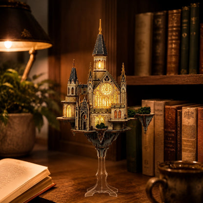 A miniature illuminated castle model displayed on a clear stand on a wooden table, surrounded by a warm lamp, books, and a mug in a cozy room setting.