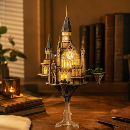 A miniature gothic castle model with warm yellow internal lighting, displayed on a clear pedestal stand on a wooden table in a cozy, dimly lit room with books and a candle in the background.