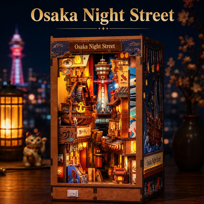 A completed Japanese Osaka Night Street DIY wooden model kit displayed on a wooden table, illuminated with warm internal lights against a dark, blurred background featuring cherry blossoms and a lantern. The words 'Osaka Night Street' are written at the top of the image.