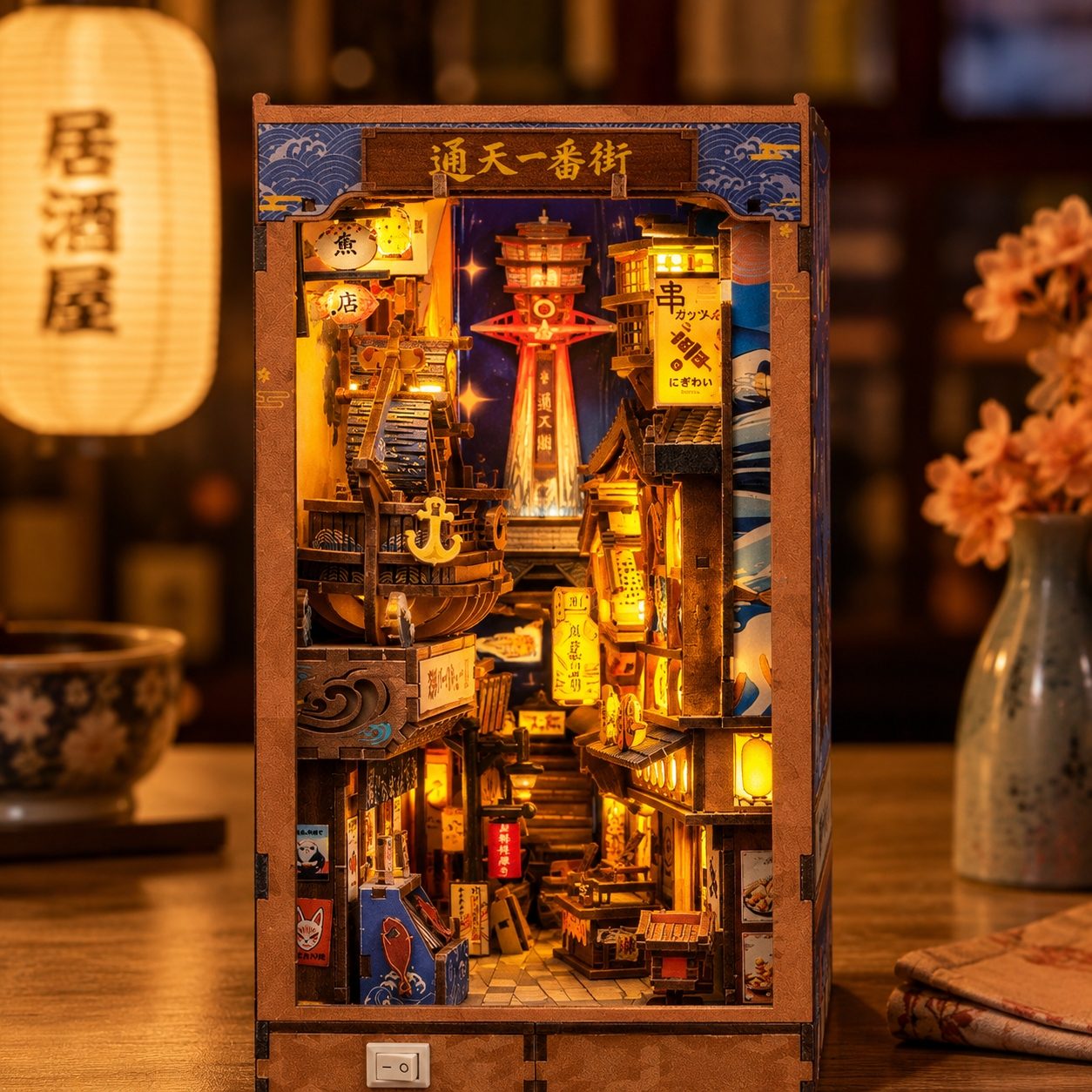 A wooden Japanese street scene DIY book nookl kit with intricate miniature details, glowing warm yellow lanterns, and a central tower, displayed on a wooden table with warm ambient background lighting.