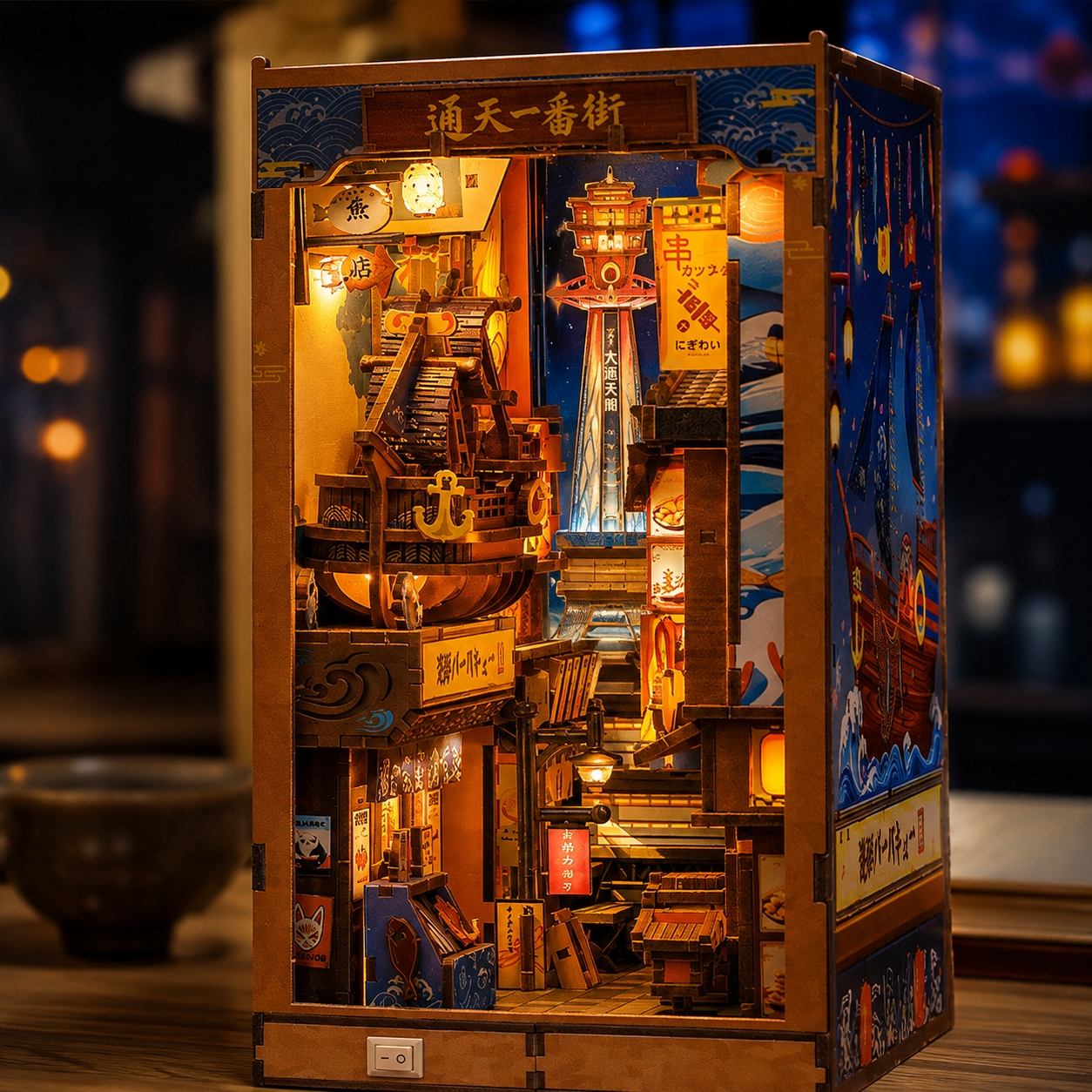 A wooden Japanese-style miniature city diorama with intricate street scenes, lanterns, and a tower, illuminated from within against a dark, softly blurred background.