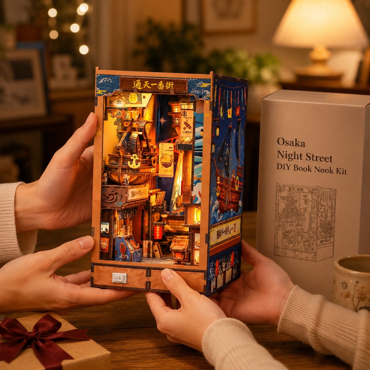 A person holds a detailed wooden Osaka Night Street DIY book nook kit, showing the illuminated interior. The product box is visible on the right, and the scene is set in a cozy, warmly lit room.