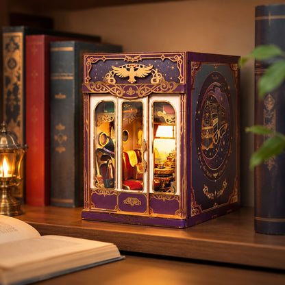 A miniature book nook style night light with a purple and gold exterior, glass-front door panels, and a warm lit interior, displayed on a wooden shelf among books.