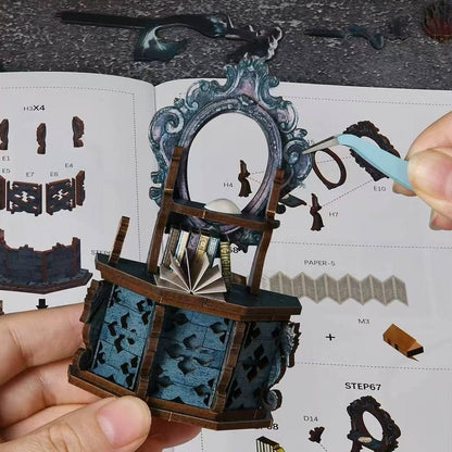 Hand holding a detailed wooden craft model of a dressing table with an open book on a table with instructions.