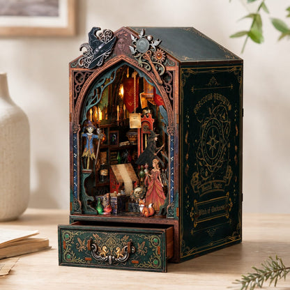 A miniature bookcase or music box with a dark green and bronze gothic design, featuring a detailed interior scene with candles, books, and small figurines, displayed on a wooden surface.