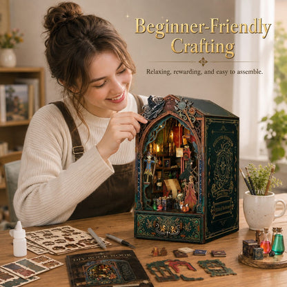 A woman smiling at a wooden table while assembling a decorative miniature book nook box. The box has an ornate dark green and gold design with a lit interior showing tiny figurines and candles.