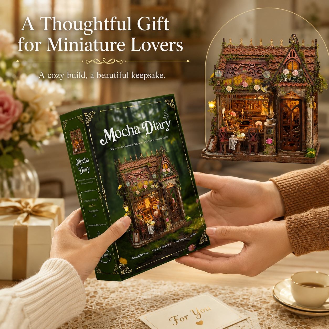 A person holds a green Mocha Diary miniature book in both hands, with a beautifully lit miniature fairy house displayed in a circular inset. The scene is set in a cozy, warm-toned room with a gift box and a cup of tea on the table, under the text 'A Thoughtful Gift for Miniature Lovers'.