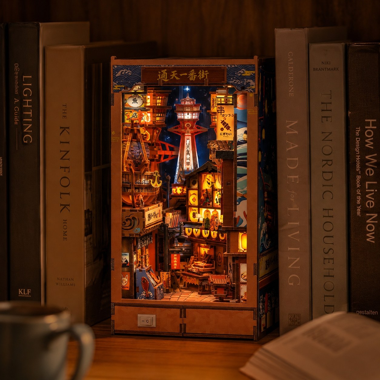 A wooden Japanese street scene DIY book nook model kit with glowing LED lights, displayed on a bookshelf between books.