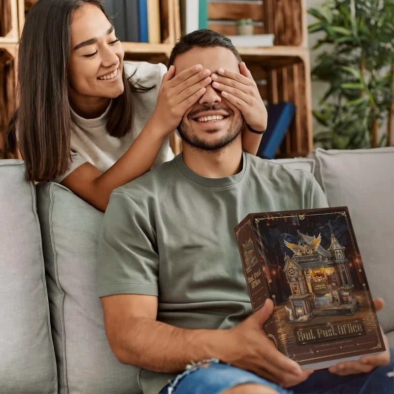 Woman covering a man's eyes with a miniature house package titled 'Owl Post Office' in a cozy living room.