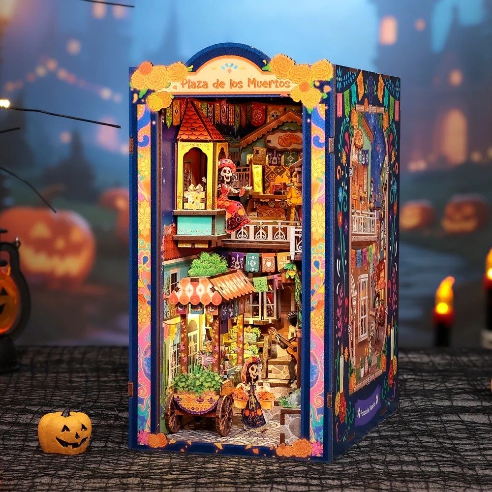 A miniature dollhouse labeled 'Plaza de los Muertos' stands lit up on a dark Halloween-themed surface. The structure is highly detailed with colorful Day of the Dead themed interiors, including a street stall, skeleton figures, and ornate blue borders.