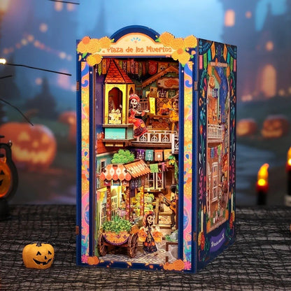A miniature dollhouse labeled 'Plaza de los Muertos' stands lit up on a dark Halloween-themed surface. The structure is highly detailed with colorful Day of the Dead themed interiors, including a street stall, skeleton figures, and ornate blue borders.