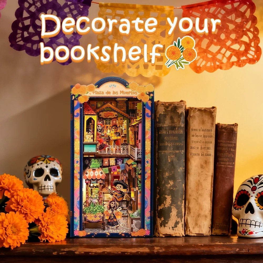 A decorative bookshelf miniature book nook featuring a colorful Day of the Dead market scene, styled on a wooden shelf next to old books, sugar skull decorations, and orange marigolds under a festive banner.