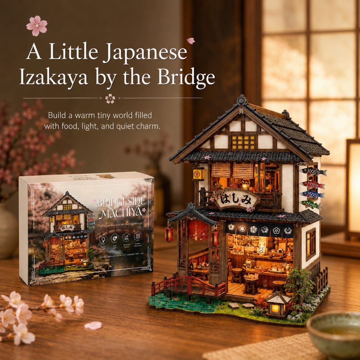 A Japanese-style izakaya miniature building kit shown with its retail box beside the assembled model. The product is photographed on a wooden table in a warm, softly lit interior setting with decorative cherry blossoms and a bowl, so this is Lifestyle rather than Product/Packaging.