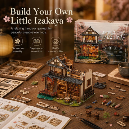 A DIY wooden building kit for a Japanese-style house labeled 'Bridgeside Machiya' is shown assembled on a wooden table, with its retail box, instruction booklet, small parts, and accessories arranged around it. Promotional text at the top reads 'Build Your Own Little Izakaya.