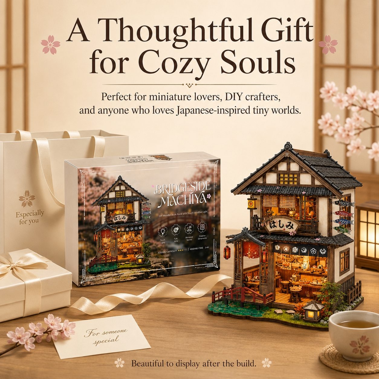 A beautifully styled product display featuring a completed miniature two-story Japanese house model with warm glowing lights, its retail box, and gift bags on a wooden table. The scene is decorated with cherry blossoms, a cup of tea, and elegant text overlays that read 'A Thoughtful Gift for Cozy Souls'.