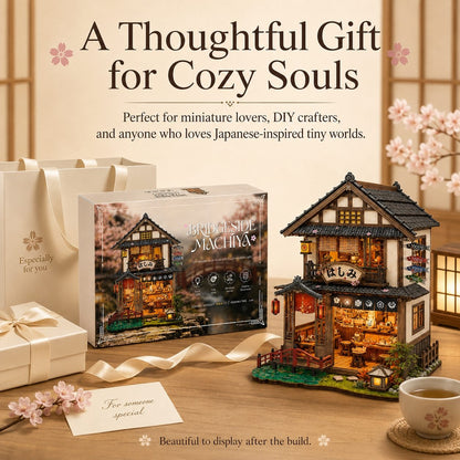 A beautifully styled product display featuring a completed miniature two-story Japanese house model with warm glowing lights, its retail box, and gift bags on a wooden table. The scene is decorated with cherry blossoms, a cup of tea, and elegant text overlays that read 'A Thoughtful Gift for Cozy Souls'.