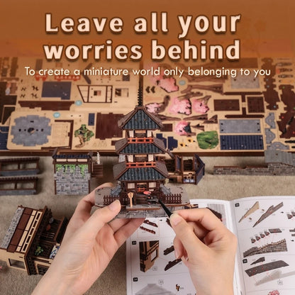 A person's hands are shown assembling a miniature Japanese pagoda model using a small tool, with an instruction booklet open below. The background displays a fully built miniature world with various wooden structures, boats, and terrain pieces, under a text overlay that reads 'Leave all your worries behind'.