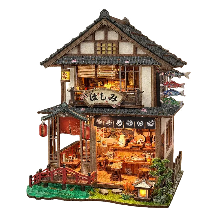 A detailed miniature Japanese-style house model with a lit interior, featuring a two-story structure with a dark tiled roof, wooden walls, a small bridge over green water, and decorative lanterns and fish flags, displayed on a wooden surface.