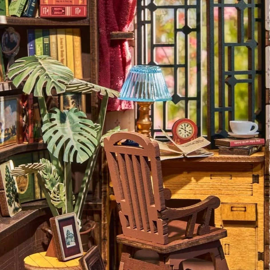 Wooden desk with chair, lamp, and books in a cozy miniature room with plants and window.