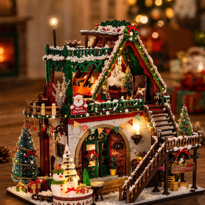 A detailed miniature Christmas house model with warm glowing lights, snow-covered roofs, a staircase, small trees, and festive decorations, photographed in a softly blurred holiday setting.