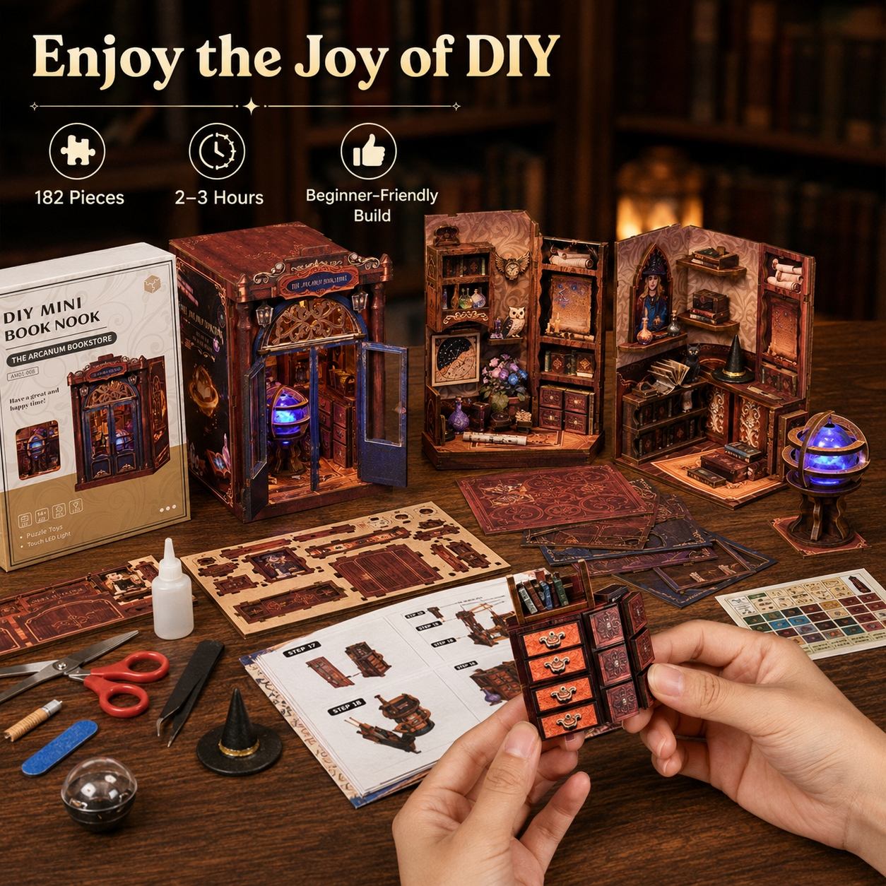 A DIY miniature book nook kit shown on a wooden table with its box, instruction sheet, tools, and assembled models. A person's hands are holding a small orange-brown book nook piece in the foreground.