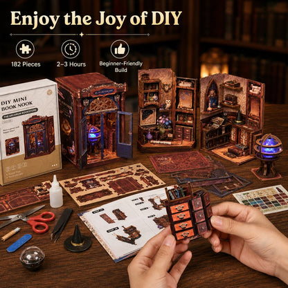 A DIY miniature book nook kit shown on a wooden table with its box, instruction sheet, tools, and assembled models. A person's hands are holding a small orange-brown book nook piece in the foreground.