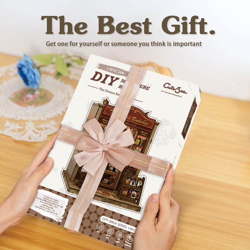 DIY book nook model kit packaging with a pink ribbon held by hands on a wooden surface.