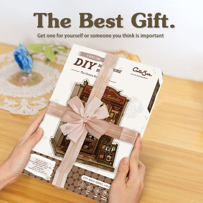 DIY book nook model kit packaging with a pink ribbon held by hands on a wooden surface.