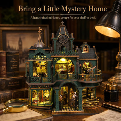 A decorative miniature detective-themed house with teal and dark gray exterior details and warm yellow interior lighting, displayed on a desk with books, a magnifying glass, ink bottle, and lamp in a cozy room setting.