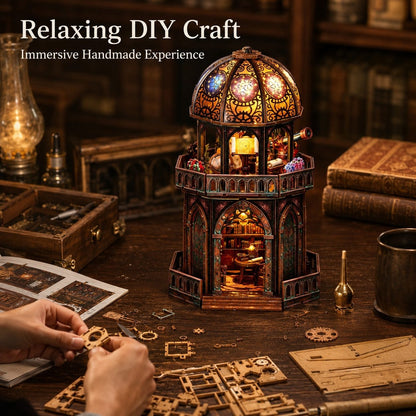 A miniature craft model resembling a stained-glass tower or observatory sits on a wooden table in a cozy, dimly lit workshop setting. Hands are visible in the lower left holding small craft pieces, with books, tools, and a brass cup around it.