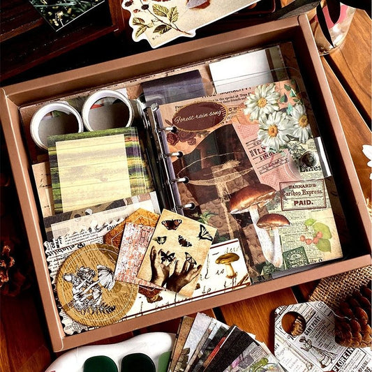 Scrapbooking supplies including papers, stickers, and a notebook in a box on a wooden surface.