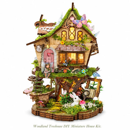 A woodland treehouse DIY miniature house kit displayed on a clean white background. The intricate fairy-tale style house features a mossy green roof, warm glowing windows, a pink fairy door, a small garden, mushrooms, butterflies, and a white rabbit figure on the base.