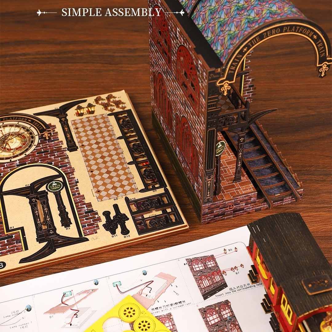 Miniature Model train station set with detailed architecture on a wooden table, including a partially assembled model, pre-cut wooden boards, and assembly manual.