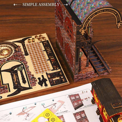 Miniature Model train station set with detailed architecture on a wooden table, including a partially assembled model, pre-cut wooden boards, and assembly manual.