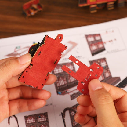 Red wooden model pieces held by hands with a blurred background of a model train set.