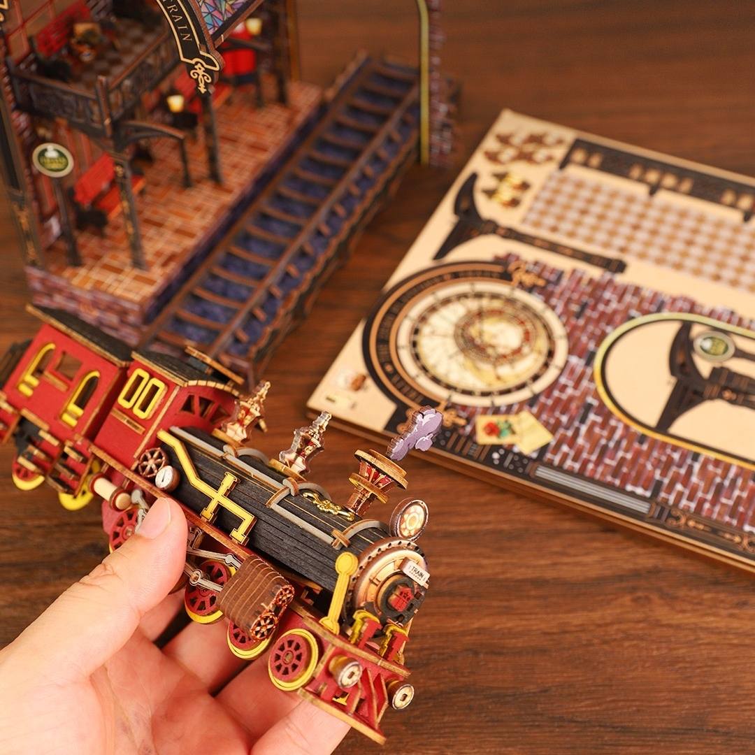 Hand holding a small miniature model train on a wooden table with a detailed model train set in the background.